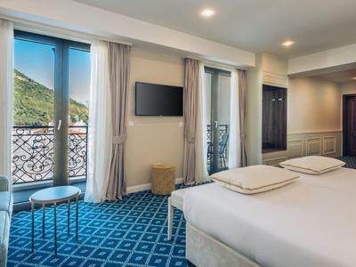 Helles Hotelzimmer mit Doppelbett, blauem Teppich und Balkon mit Blick auf Berge.