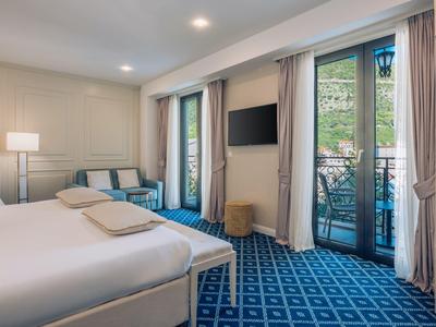 Hell eingerichtetes Hotelzimmer mit Doppelbett, blauem Teppich und Balkon mit Blick ins Grüne.