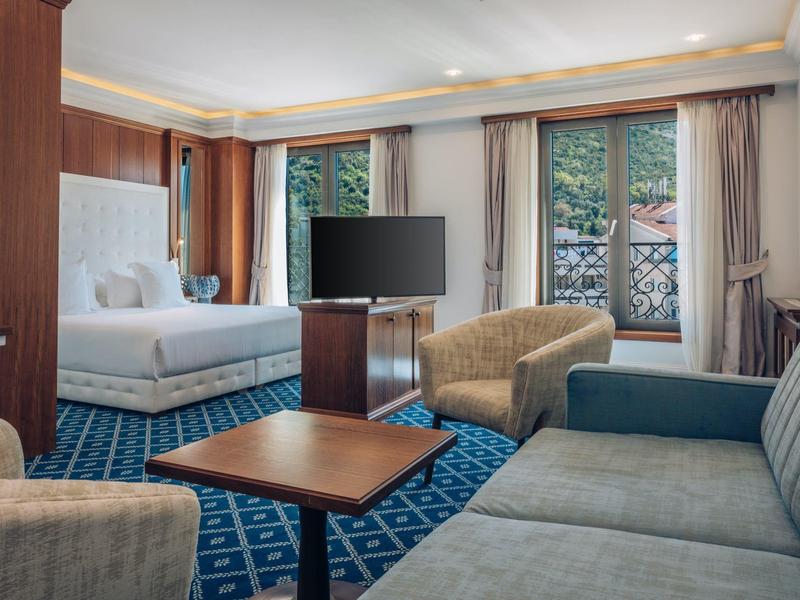 Helles Hotelzimmer mit Doppelbett, Sofas und großem Fenster mit Blick auf Bäume.