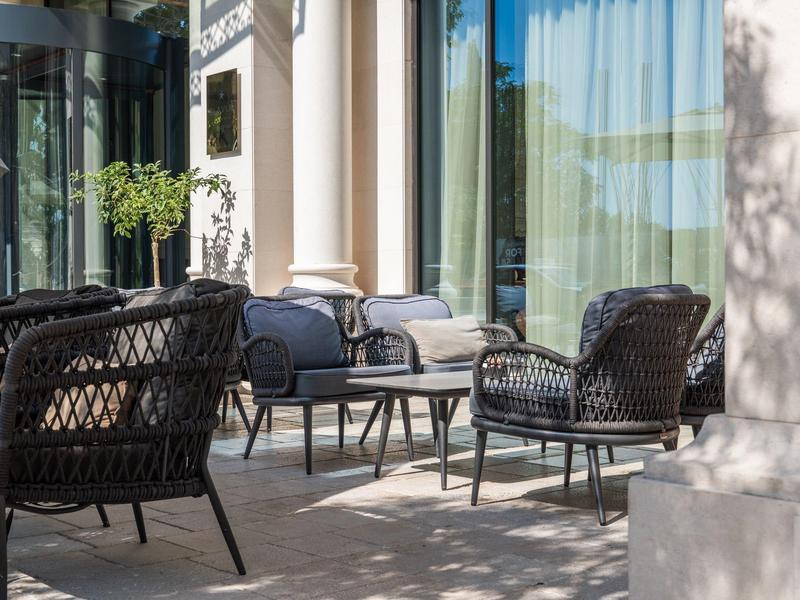 Gemütliche Sitzgruppe mit schwarzen Korbstühlen und Tischen auf einer Terrasse vor einem Hotel.