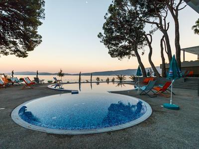 Runder Pool mit Liegestühlen und Bäumen am Meer bei Sonnenuntergang.