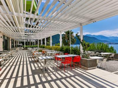 Hotelterrasse mit Schattenmuster, roten Stühlen und Blick auf See und Berge.