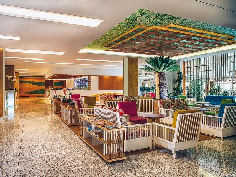 Bright hotel lobby with seating area, patterned ceiling, and large window with plants.
