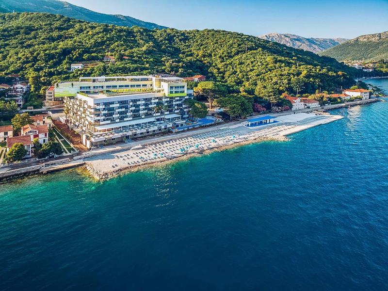 Hotel by the sea with sandy beach and sun loungers, surrounded by wooded hills.
