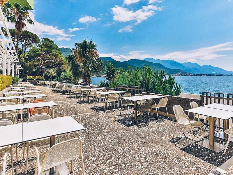Hotel terrace with tables and chairs, sea view, and mountains in the background.