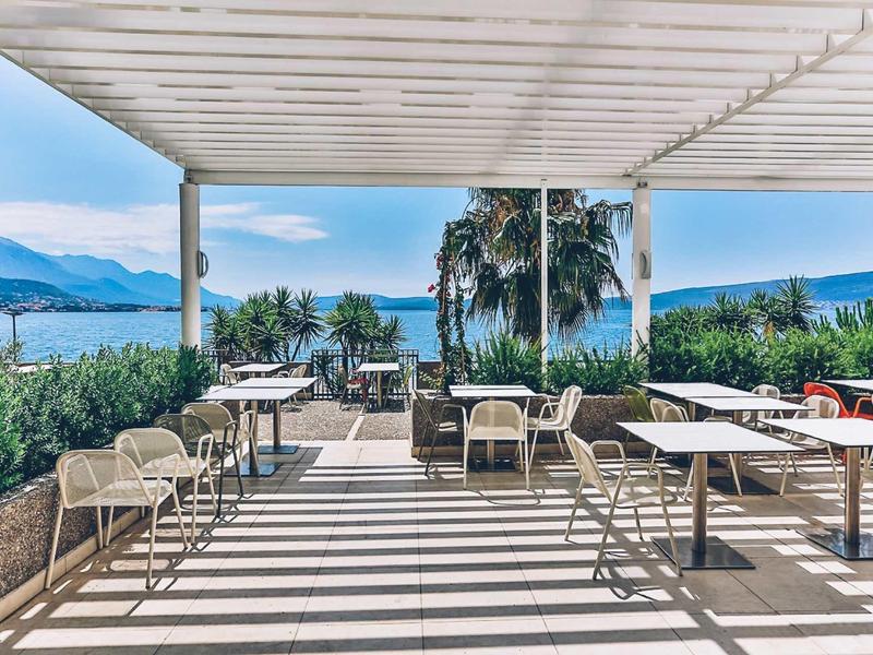 Outdoor terrace with tables and chairs overlooking the sea and palm trees.