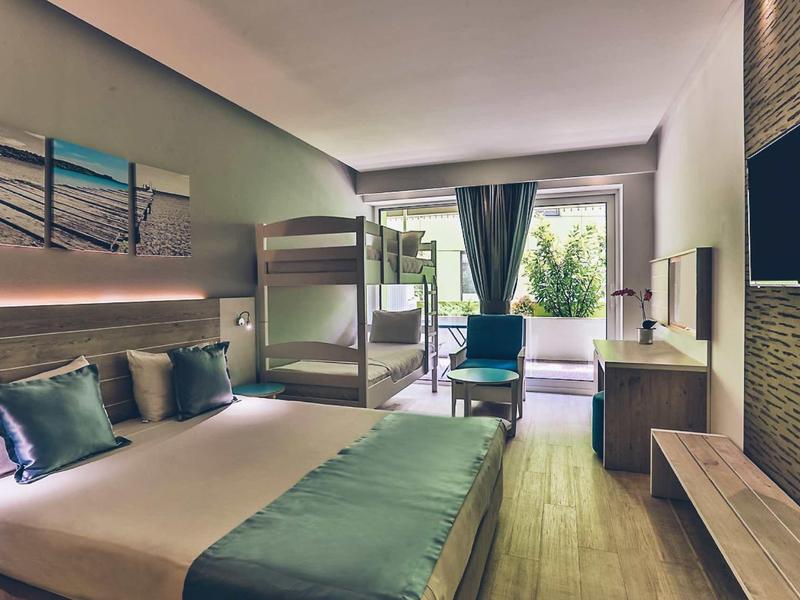 Modern hotel room with double bed, bunk bed, and balcony overlooking greenery.