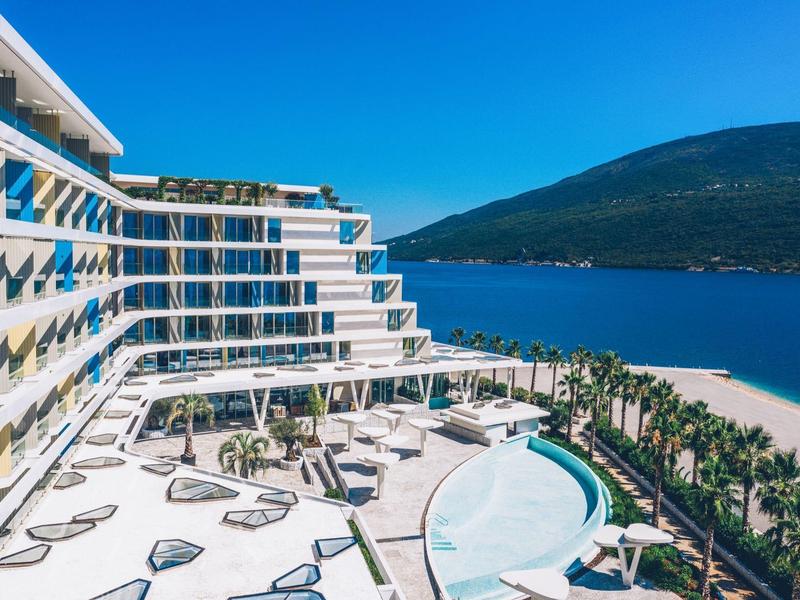Modern hotel with pool, palm trees, and view of blue sea and mountains under clear sky.
