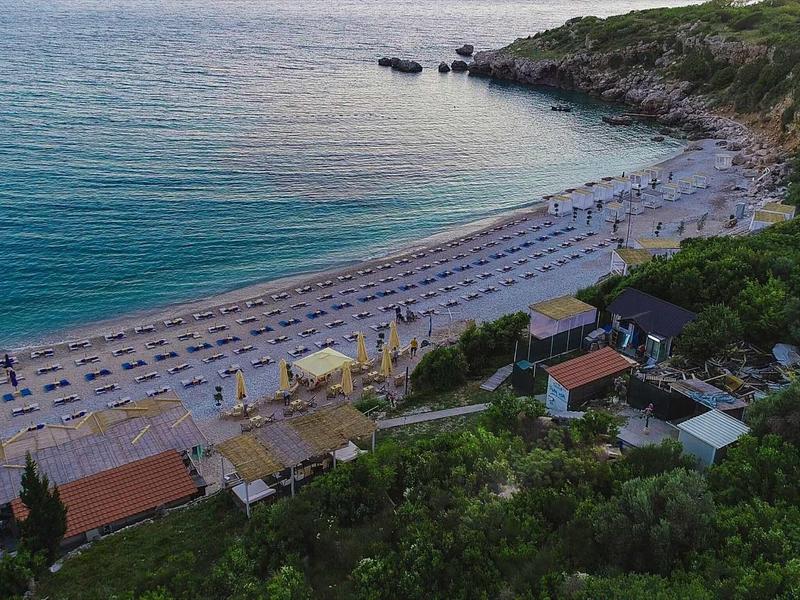 Langer Sandstrand mit Sonnenliegen, klarem blauem Meer und bewaldeten Hügeln im Hintergrund.