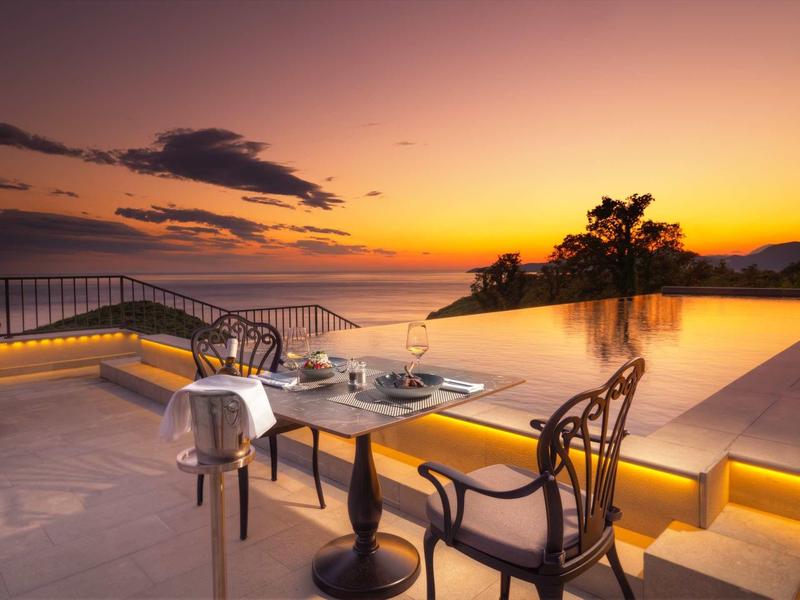 Abendstimmung an einem beleuchteten Pool mit Tisch und zwei Stühlen, Blick auf Hügel und Sonnenuntergang.