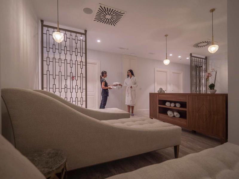 Moderne, helle Wellness-Lounge mit gemütlichen Liegen, Holztheke und zwei Personen im Gespräch.