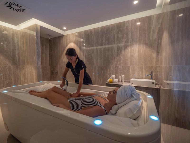 Frau liegt entspannt in Weißer Badewanne, andere Person massiert ihren Rücken, warme Beleuchtung.