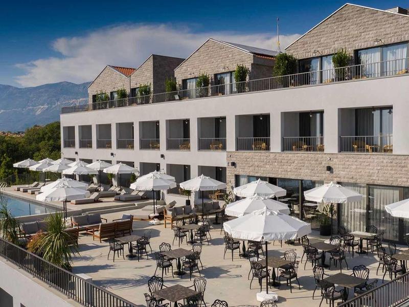 Modernes Hotel mit großer Terrasse, Sitzgelegenheiten, Sonnenschirmen und Bergpanorama im Hintergrund.