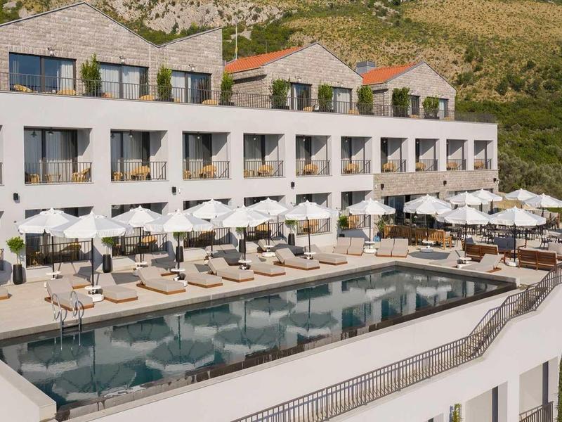 Modernes Hotel mit Pool, Liegestühlen und weißen Sonnenschirmen vor grünen Hügeln.
