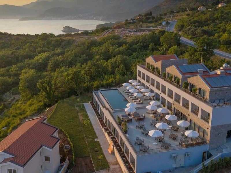 Hotelgebäude mit Terrassen, weißen Sonnenschirmen an einem Hang nahe Wald und Wasser.