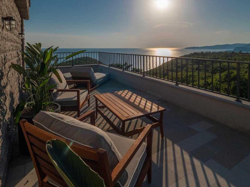 Terrasse mit Holzmöbeln, Pflanzen und Meerblick bei Sonnenuntergang, balustradenförmig begrenzt.