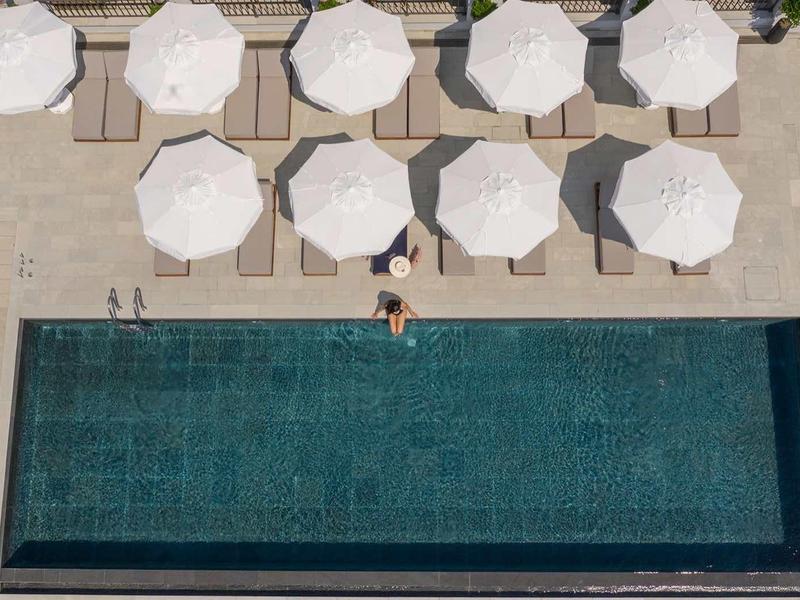 Schwarzer rechteckiger Pool mit Menschen und weißen Sonnenschirmen auf beigem Steinboden.
