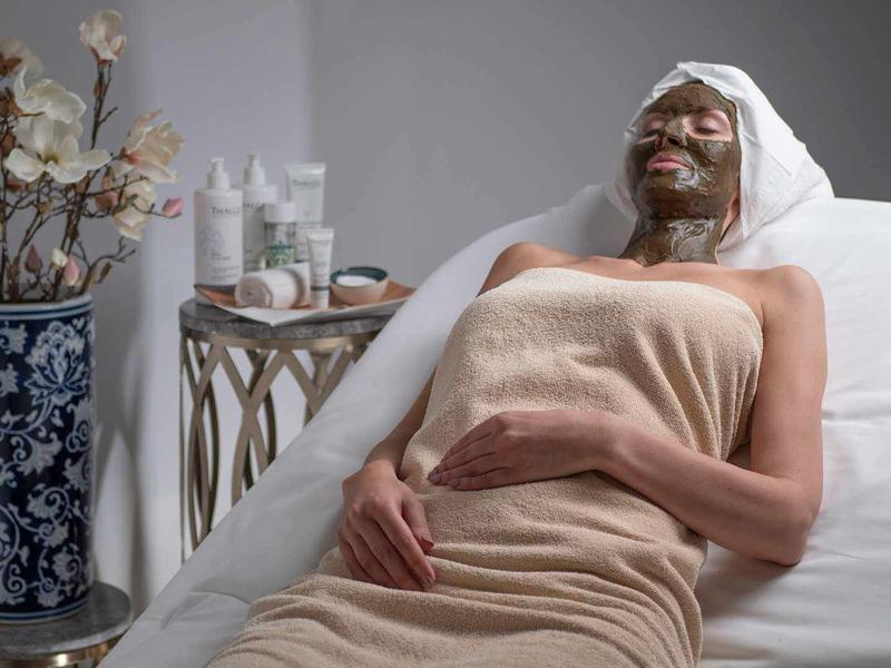 Ältere Frau entspannt in Spa mit Gesichtsmaske und Handtuch auf Liege, neben Blumenvase und Pflegeprodukten.