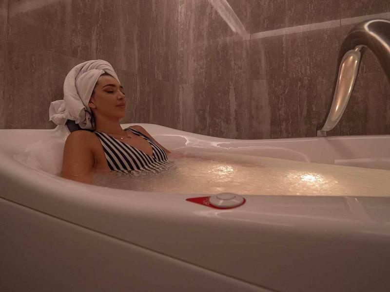 Frau entspannt in moderner Badewanne mit Duschkopf, Handtuch auf dem Kopf und beigem Licht.