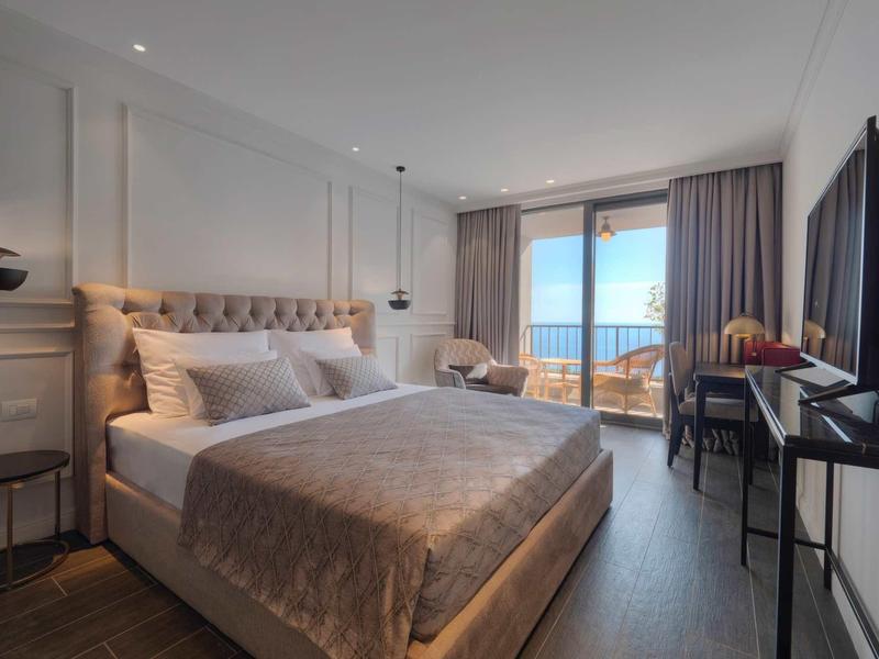 Modernes Schlafzimmer mit großem Bett, Meerblick und Balkon, weichen Vorhängen und TV an der Wand.