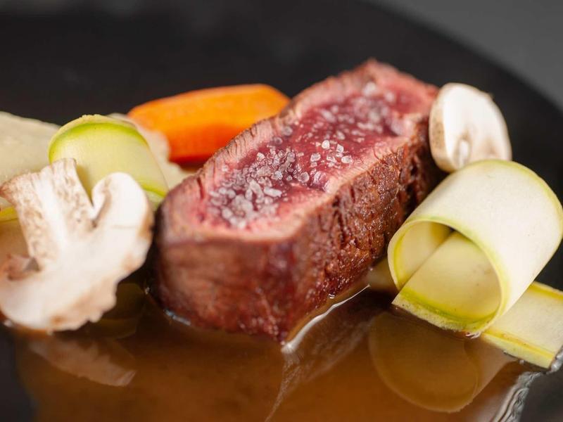 Saftiges Stück Rindfleisch auf dunklem Teller mit Karotten, Pilzen, Lauch und Sauce.