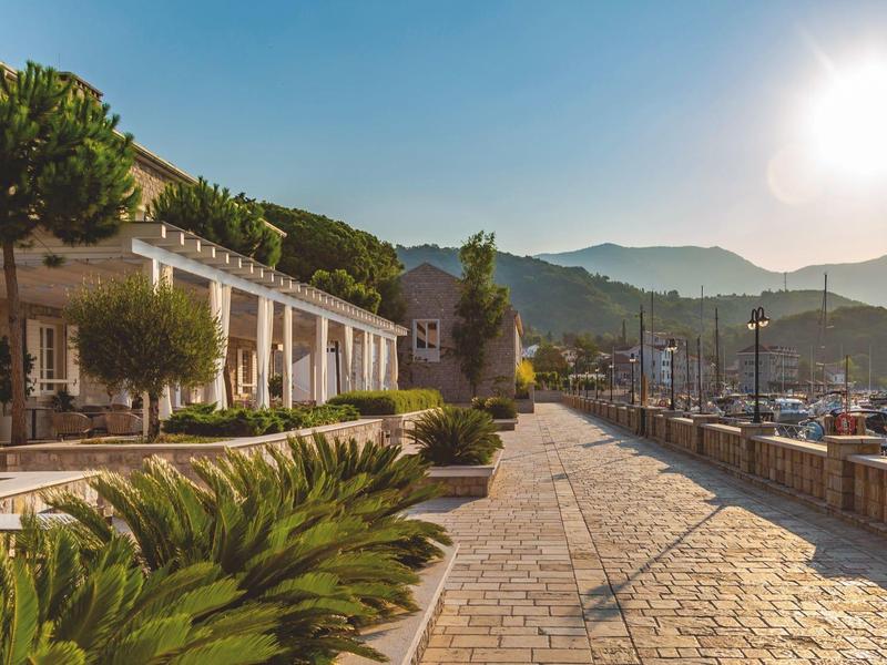 Promenade portuaire ensoleillée avec bâtiments, plantes et montagnes en arrière-plan.