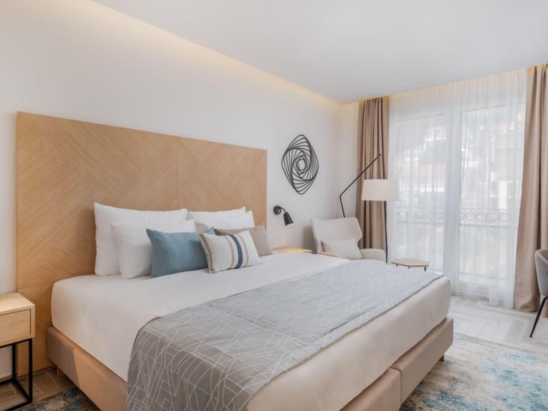 Chambre d'hôtel lumineuse avec grand lit, fauteuils confortables et grandes fenêtres avec rideaux.