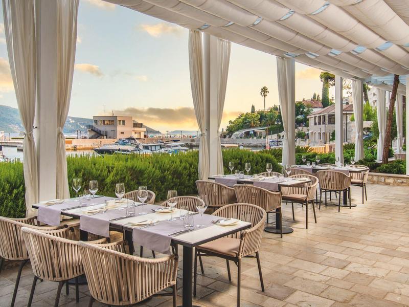 Terrasse couverte avec tables dressées offrant une vue sur le port et les montagnes au coucher du soleil.