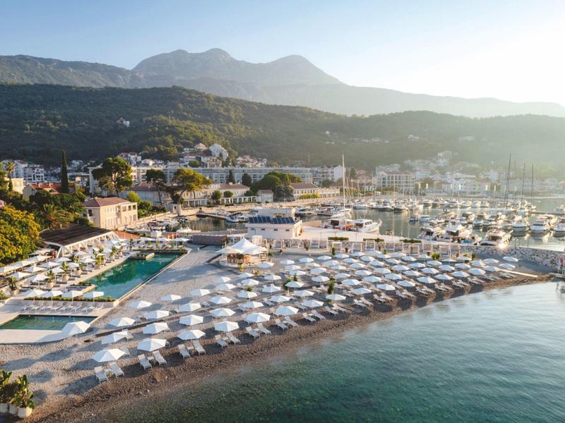 Vue d'ensemble d'une plage avec de nombreux transats et parasols blancs à côté d'un port de plaisance devant des montagnes.