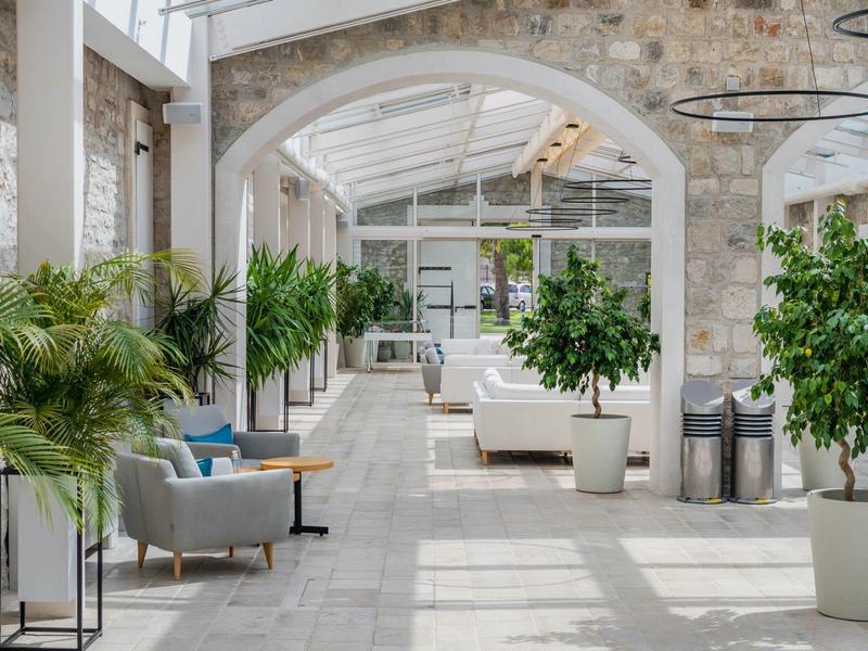 Atrium lumineux avec murs en pierre, grandes plantes et fauteuils confortables sous un toit en verre.