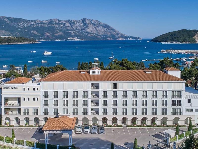 Großes weißes Gebäude mit roten Dächern am Meer, dahinter Berge und mehrere Boote auf blauem Wasser.