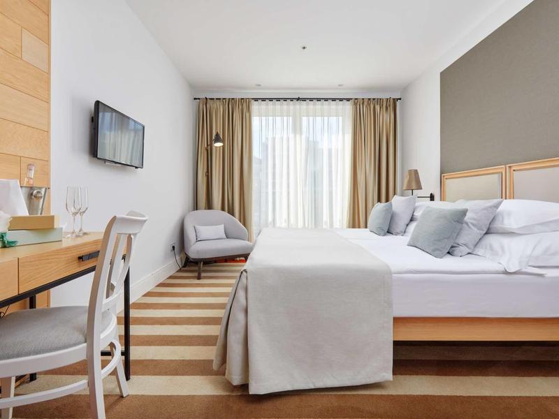 Modern eingerichtetes Hotelzimmer mit großem Bett, Schreibtisch, Fernseher und Fenster mit Gardinen.