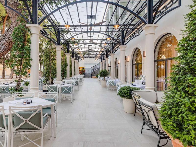 Langer überdachter Außenbereich mit weißen Tischen, Stühlen und großen Fenstern an Säulenwand.