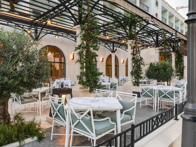 Außenbereich eines Cafés mit weißen Tischen und Stühlen, Pflanzen und einer Pergola mit Glühbirnen.