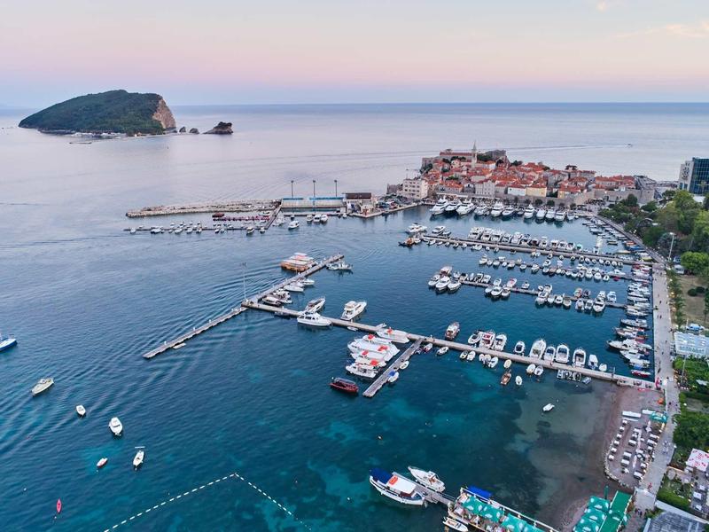 Blick auf eine Küstenstadt mit Hafen, vielen Booten und klarem, blauem Meer bei Abendlicht.
