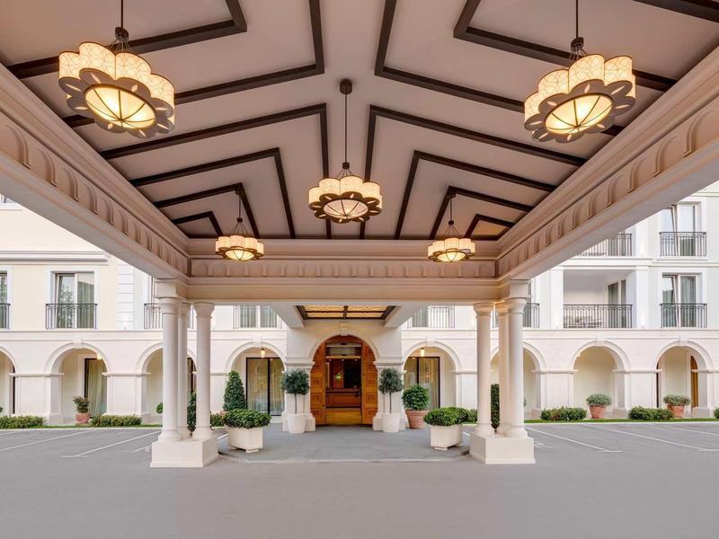 Eingangsbereich eines Gebäudes mit weißer Säulenhalle, Laternenlampen und bogenförmigen Fenstern.