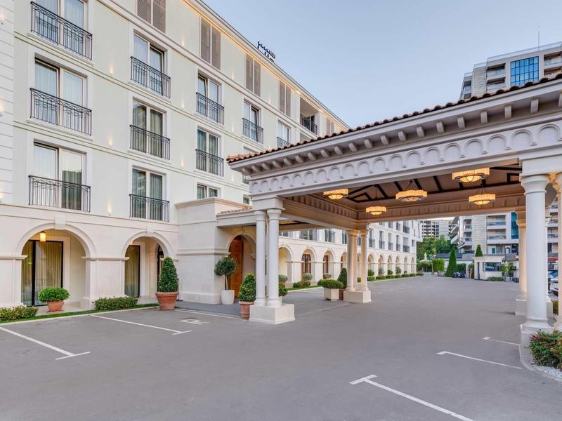 Großes, helles Gebäude mit Arkaden und überdachtem Eingangsbereich, Parkplätze im Vordergrund.