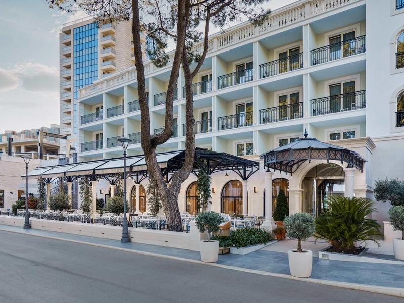 Mehrstöckiges weißes Hotelgebäude mit blauen Akzenten und Außenterrasse an Straße mit Bäumen.