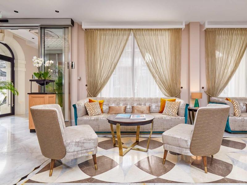 Heller, eleganter Salon mit runden Sesseln, goldfarbenem Couchtisch und großen Fenstern mit Vorhängen.