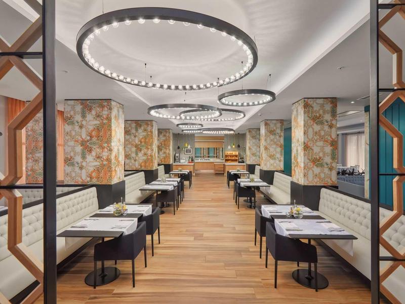 Moderner Speiseraum mit Holzfußboden, weißen Tischen, schwarzen Stühlen und großen runden Deckenleuchten.