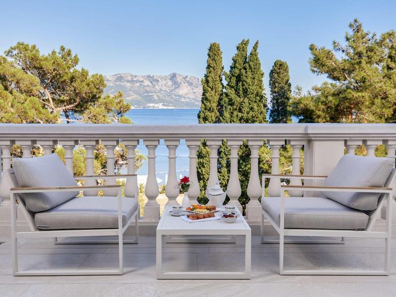 Terrassenbereich mit zwei weißen Sesseln, kleinem Tisch, Blick auf Meer und Berge im Hintergrund.