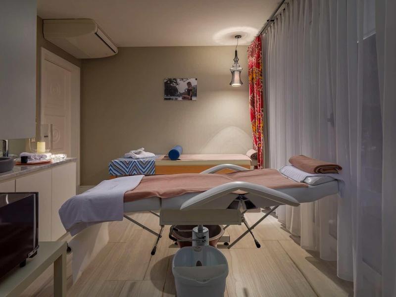 Behandlungsraum mit Massageliege, weichen Kissen, dezenter Beleuchtung und Gardinen.