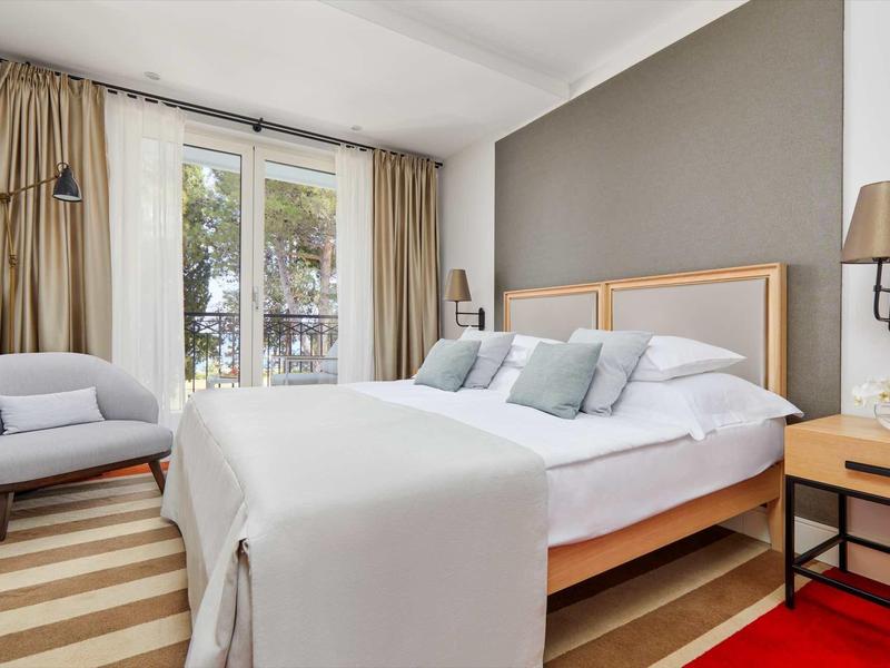 Modernes Schlafzimmer mit großem Bett, grauer Akzentwand, Sessel und Glasschiebetür zum Balkon.