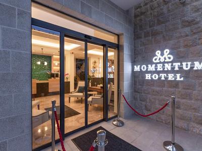 Ingresso dell'Hotel Momentum con porte scorrevoli in vetro e logo illuminato sulla parete di pietra.