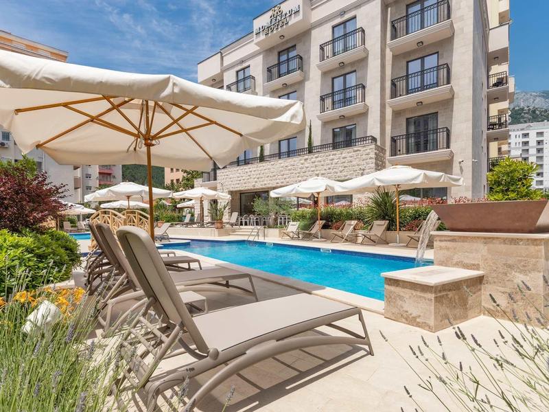Outdoor-Pool mit Liegestühlen und Sonnenschirmen vor einem mehrstöckigen Hotelgebäude bei Sonnenschein.