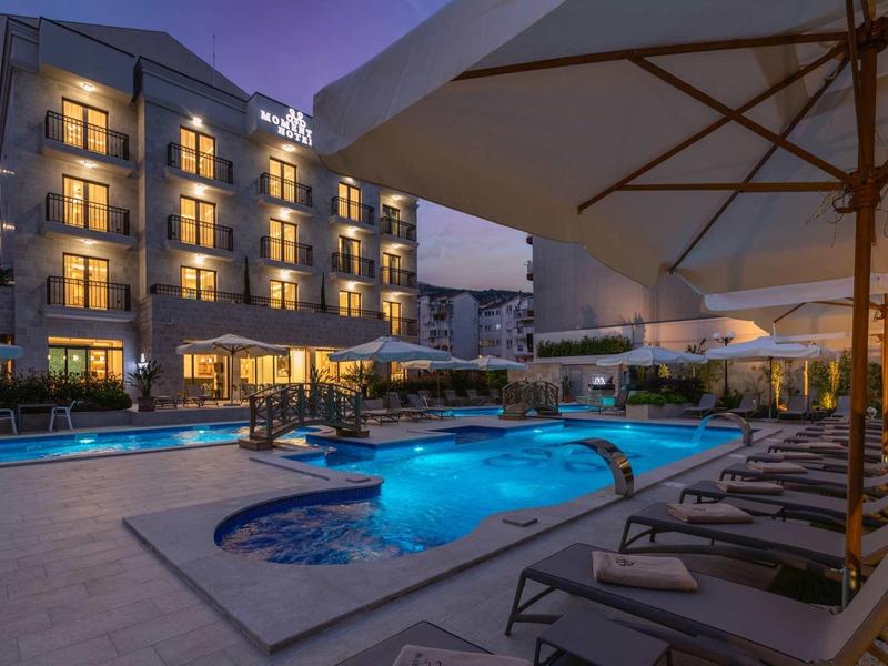 Hotel mit beleuchtetem Pool und Liegestühlen bei Abenddämmerung, großer Sonnenschirm im Vordergrund.