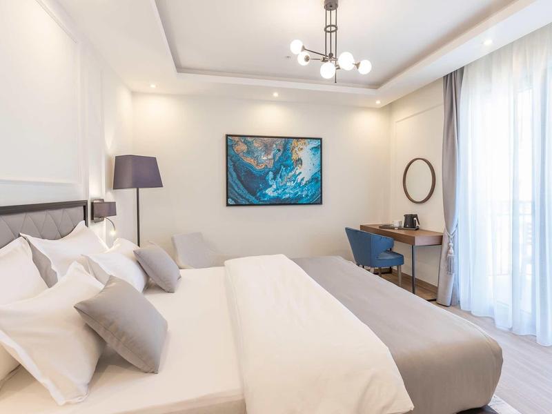 Modernes Schlafzimmer mit großem Bett, grauen Kissen, Schreibtisch und hellem Tageslicht durch Fenster.