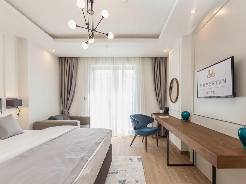 Modern eingerichtetes Schlafzimmer mit Doppelbett, Schreibtisch, Sesseln und großem Fenster mit Vorhängen.