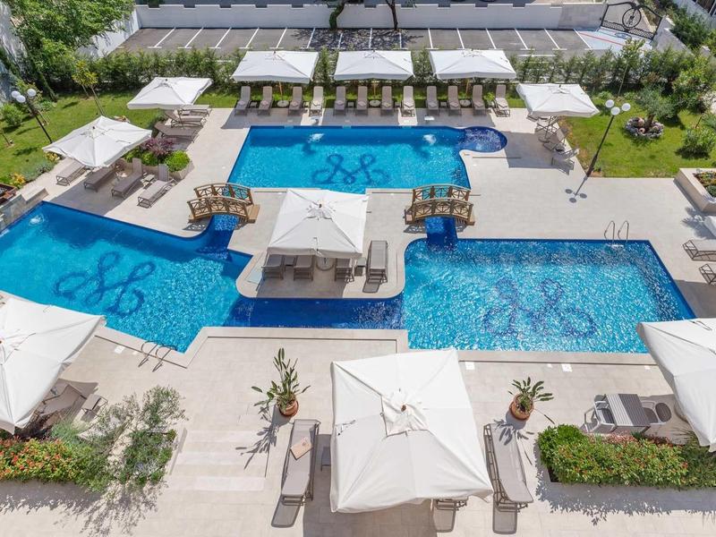 Moderner Außenpool mit klarem Wasser, weißen Liegen, Sonnenschirmen und grüner Umgebung.