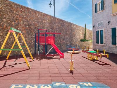 Een zonnige, lege speelplaats met glijbaan en wip naast een stenen muur en gebouw